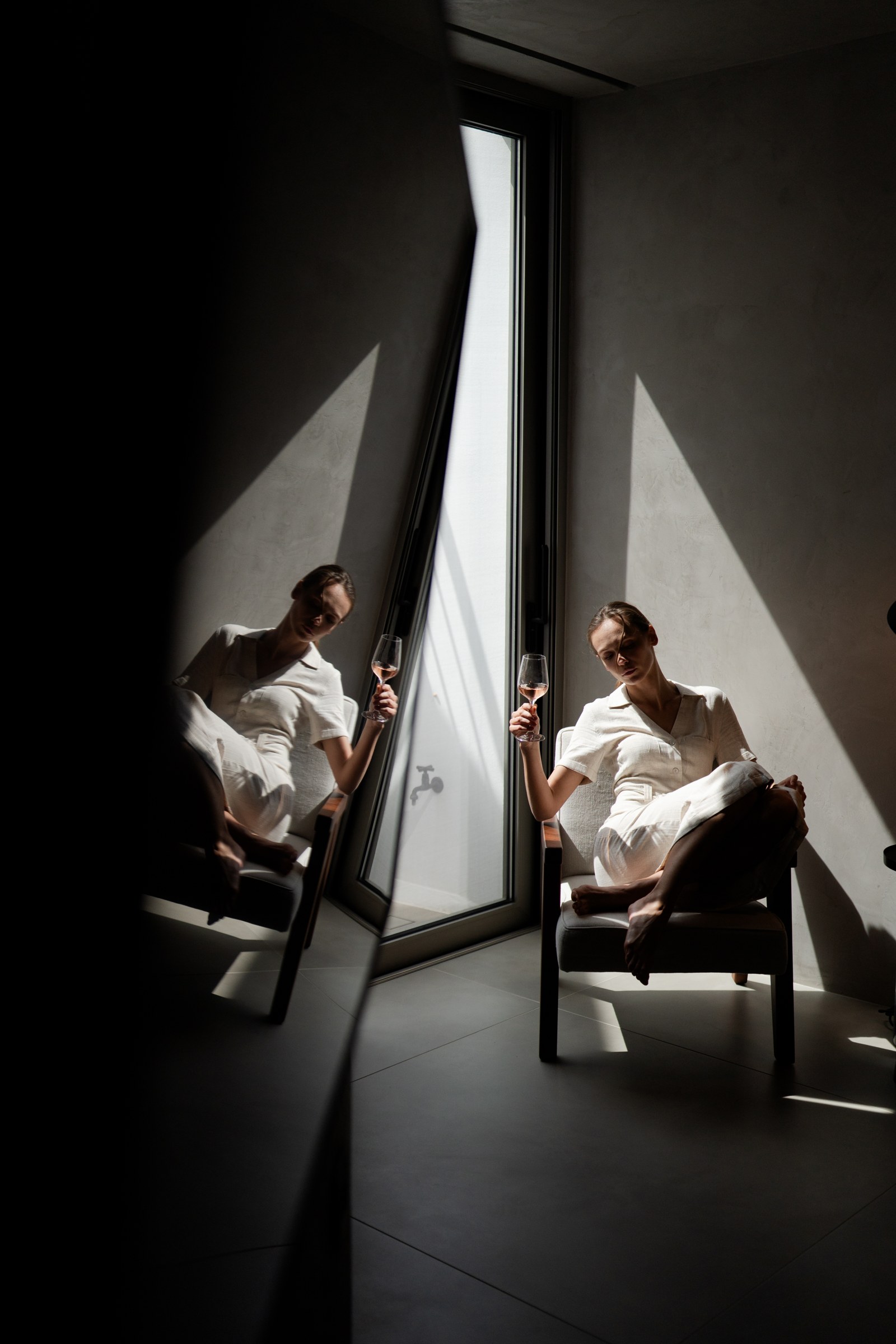 PNOES Tinos: a woman with a wine glass beside a sunlit window, reflected in a leaning mirror